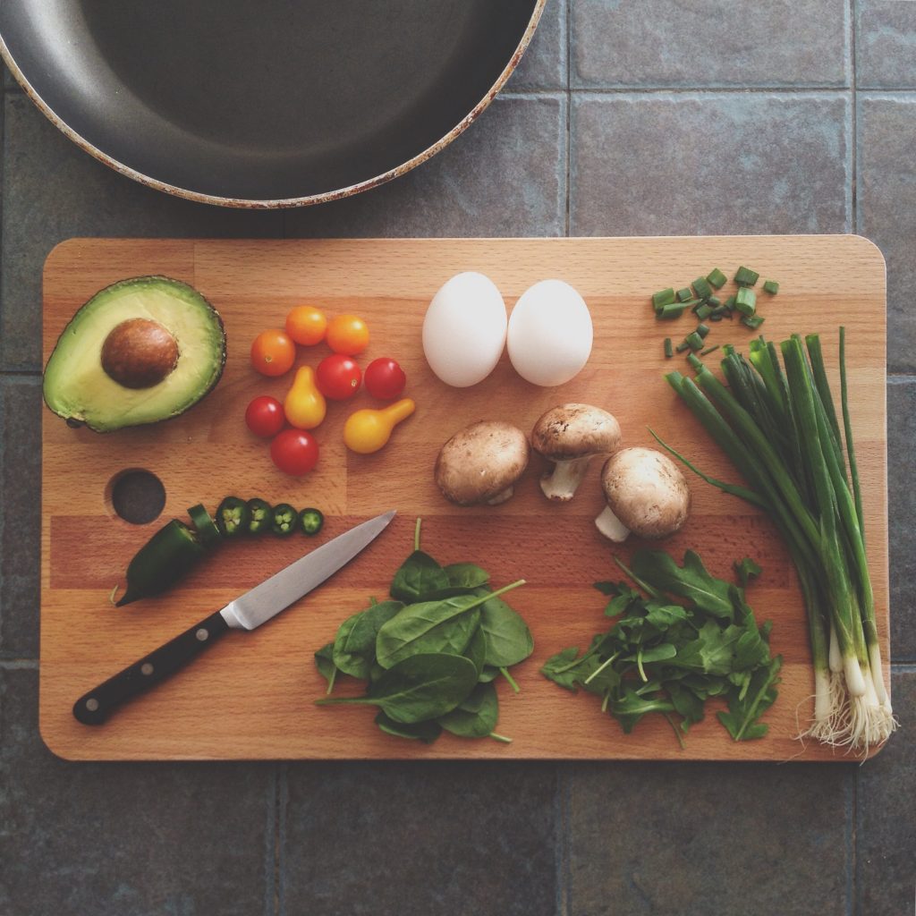 Wooden chopping board with various foods laid out, eggs, avocado, mushroom, tomatoes, herbs.